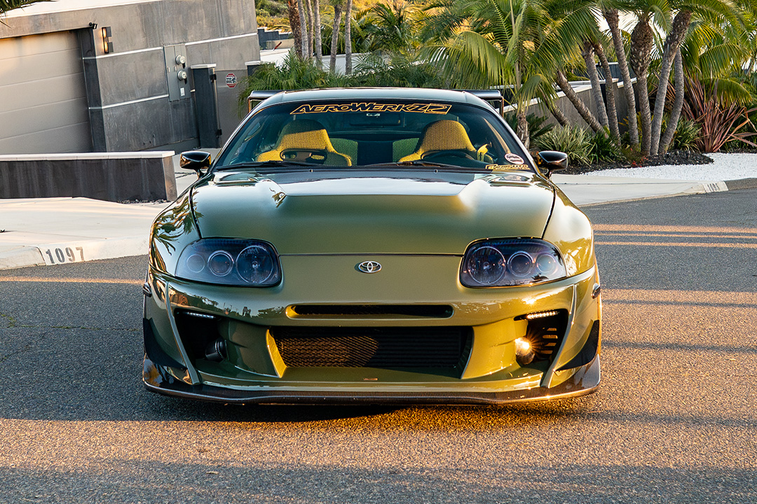Front view of Leopold Barrit’s Military Olive Green 1995 Toyota Supra parked on a palm tree-lined street at sunset.