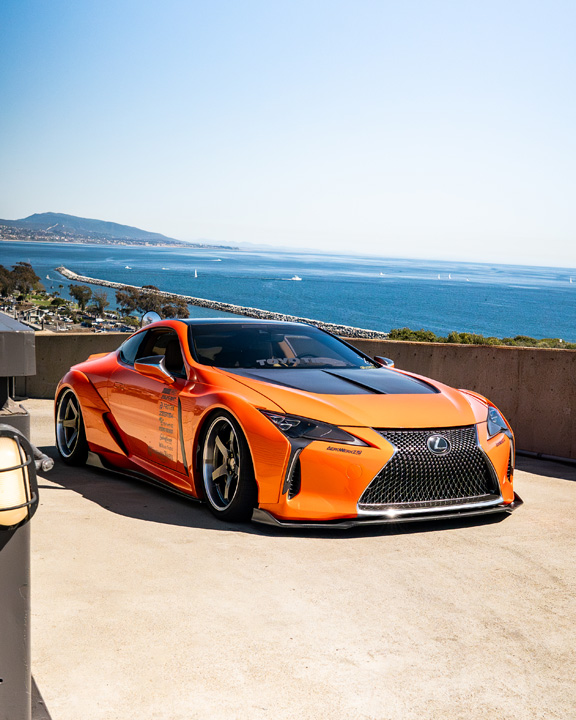 Cassandra Balthasar’s orange widebody Lexus LC500 parked above the ocean in a front three-quarter view.
