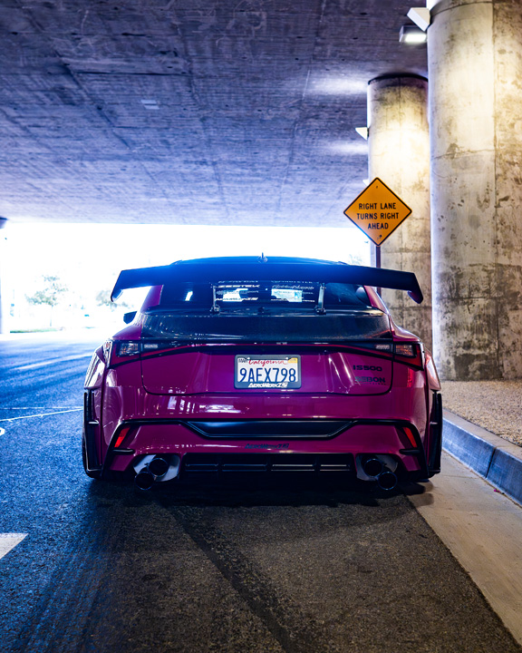 Rear view of Giuseppe Balthasar’s pink widebody Lexus IS 500 under a concrete overpass showing the large rear wing and quad exhaust.