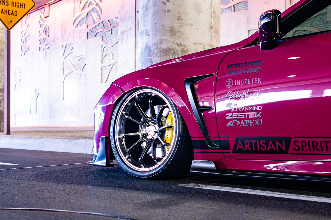 Front wheel and sponsor detail on Giuseppe Balthasar’s pink widebody Lexus IS 500 under an overpass.