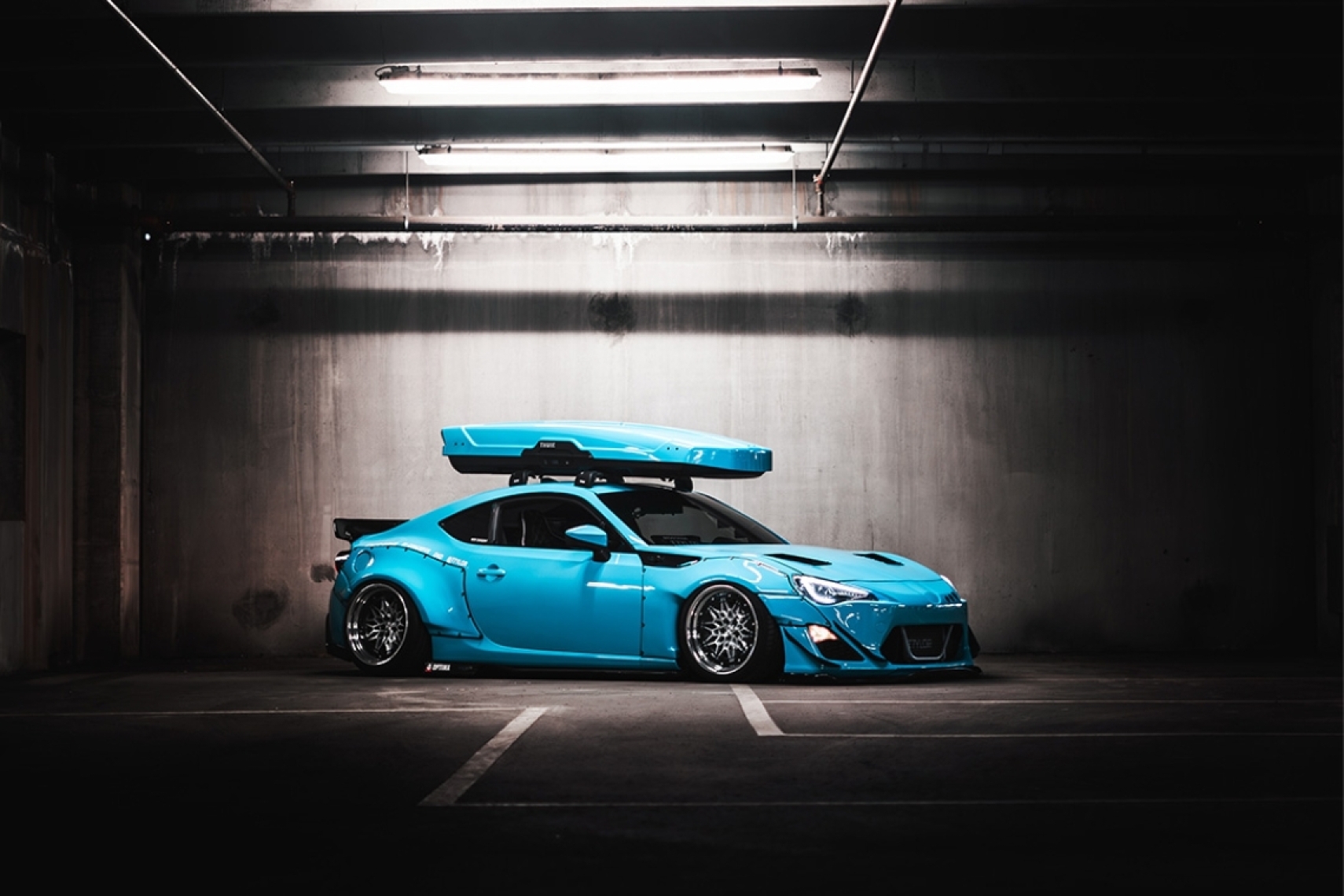 Widebody 2016 Scion FR-S with a Rocket Bunny V2 kit and roof-mounted ski box parked inside a dimly lit parking garage.