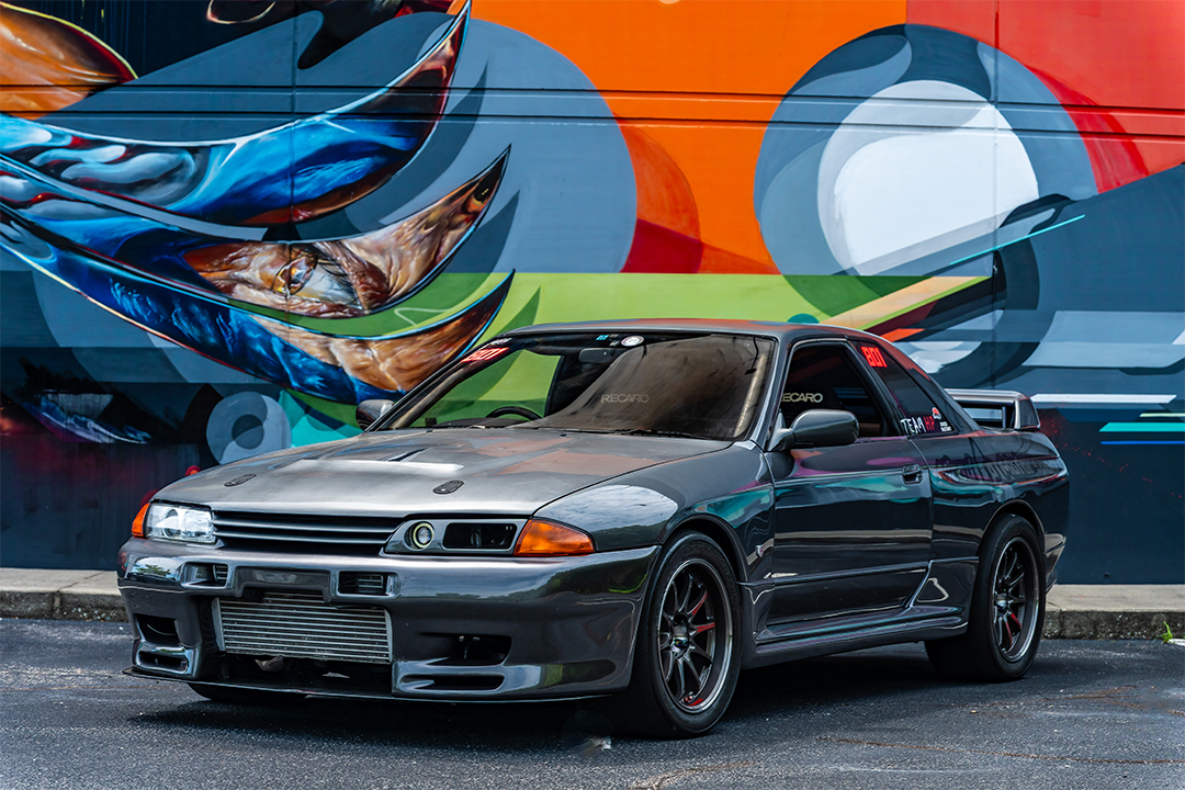 Front three-quarter view of Cee Vann’s gray 1990 Nissan Skyline GT-R parked in front of a colorful mural with exposed front-mount intercooler and black wheels.
