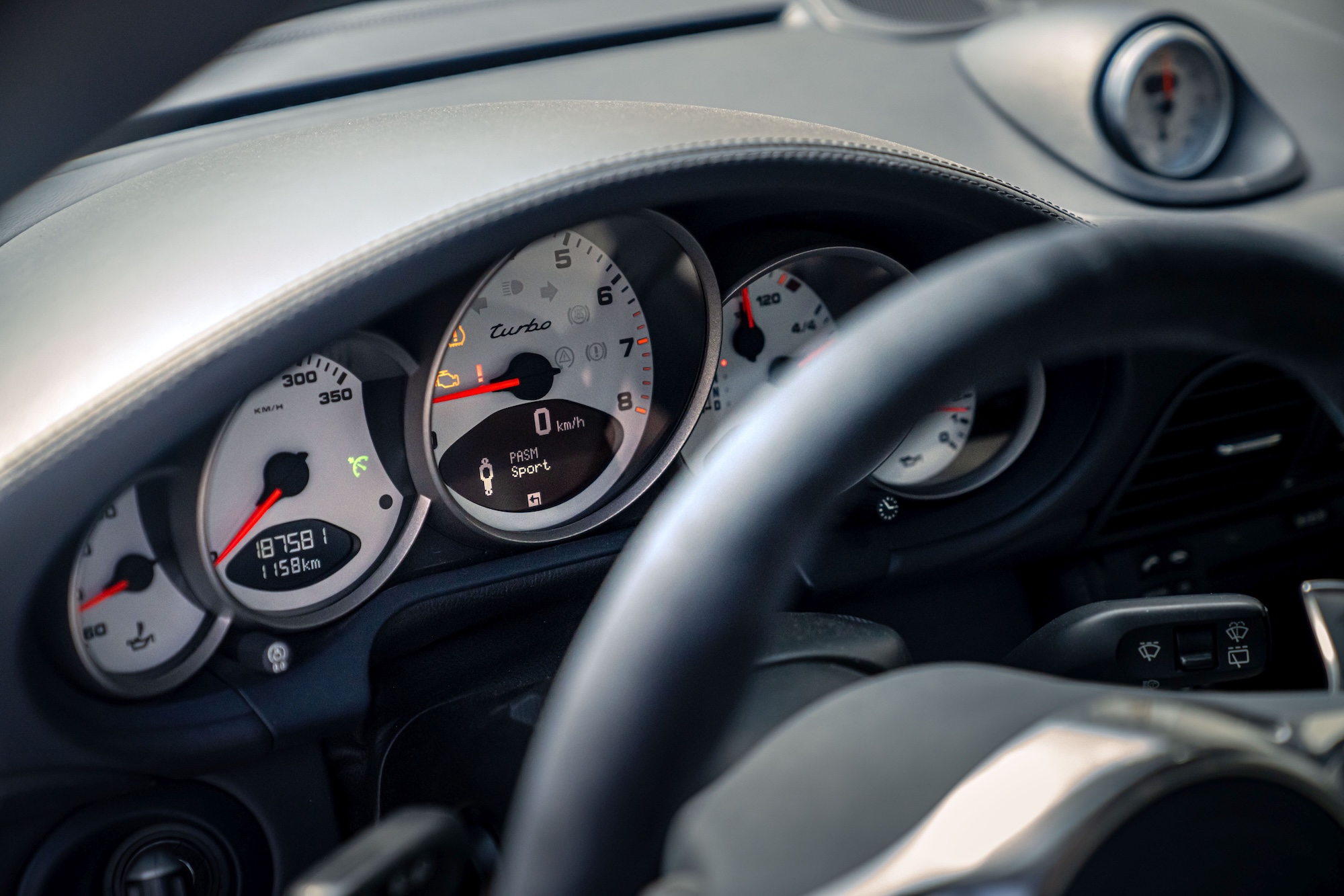 Porsche 911 997 instrument cluster showing PASM Sport mode with factory gauges and steering wheel in the foreground.