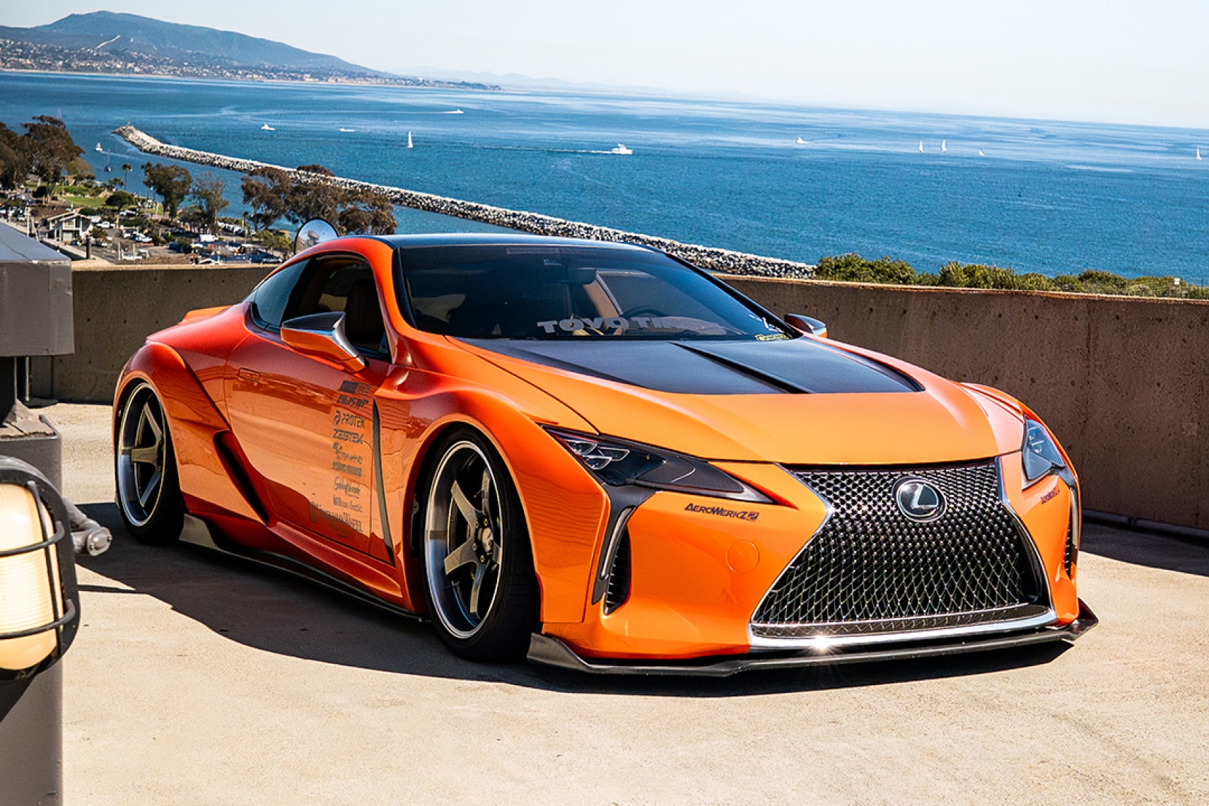 Cassandra Balthasar’s orange widebody Lexus LC500 parked above the ocean in a front three-quarter view.