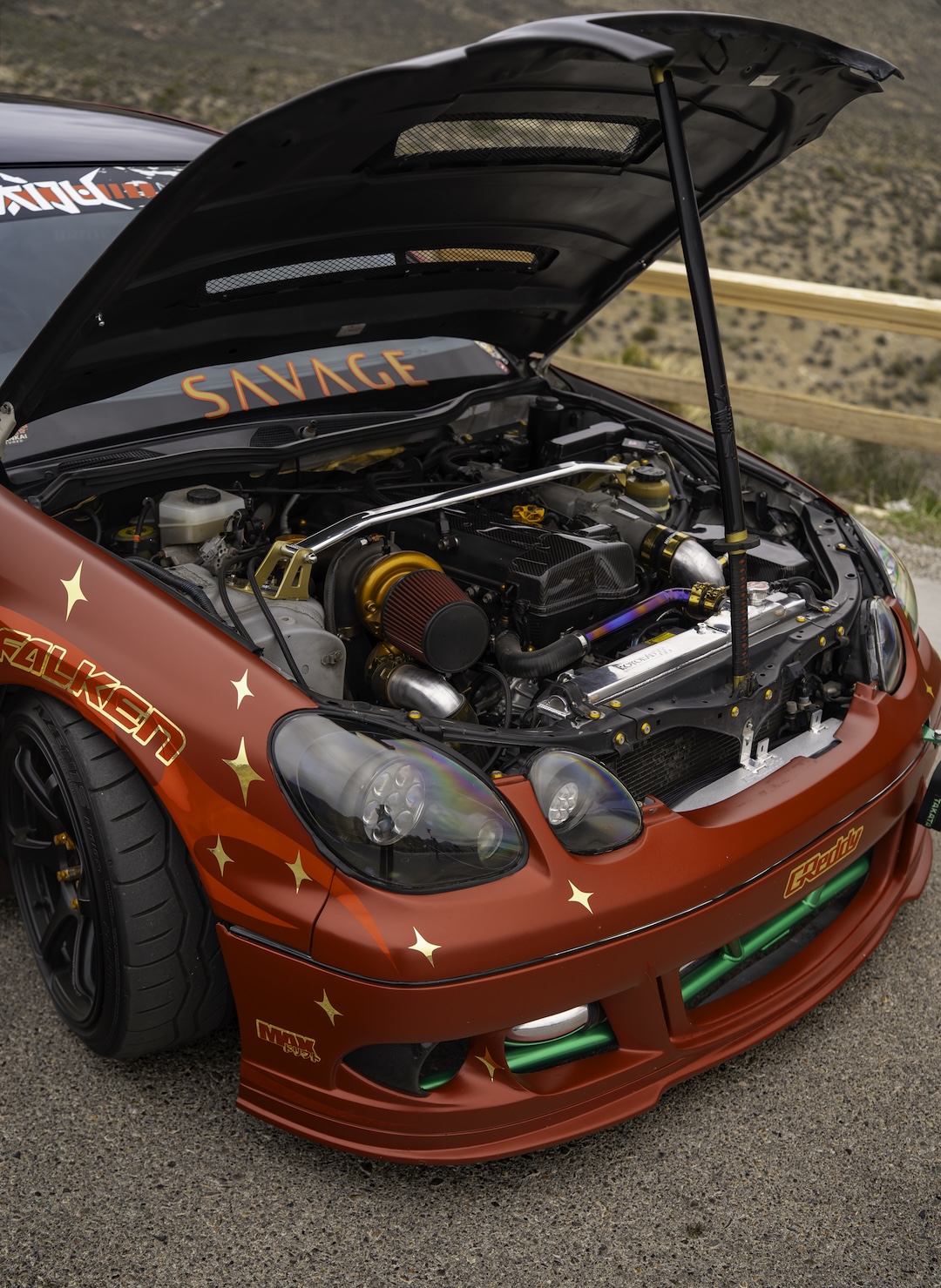 Engine bay view of Chris Burch’s satin red 1997 Toyota Aristo showing the turbocharged 2JZ setup, carbon hood, front strut bar, exposed bash bar, and custom engine bay hardware.