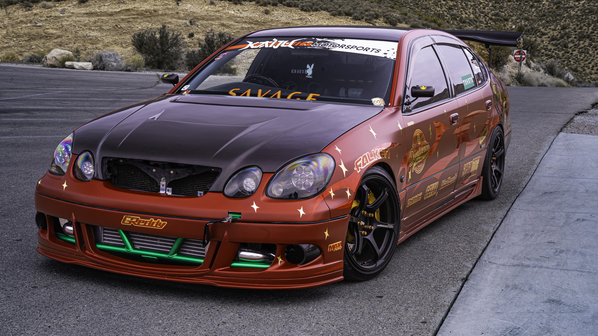 Front three-quarter view of Chris Burch’s satin red 1997 Toyota Aristo showing the RAW livery, carbon hood, exposed front bash bar, Advan RG4 wheels, and mountain backdrop.