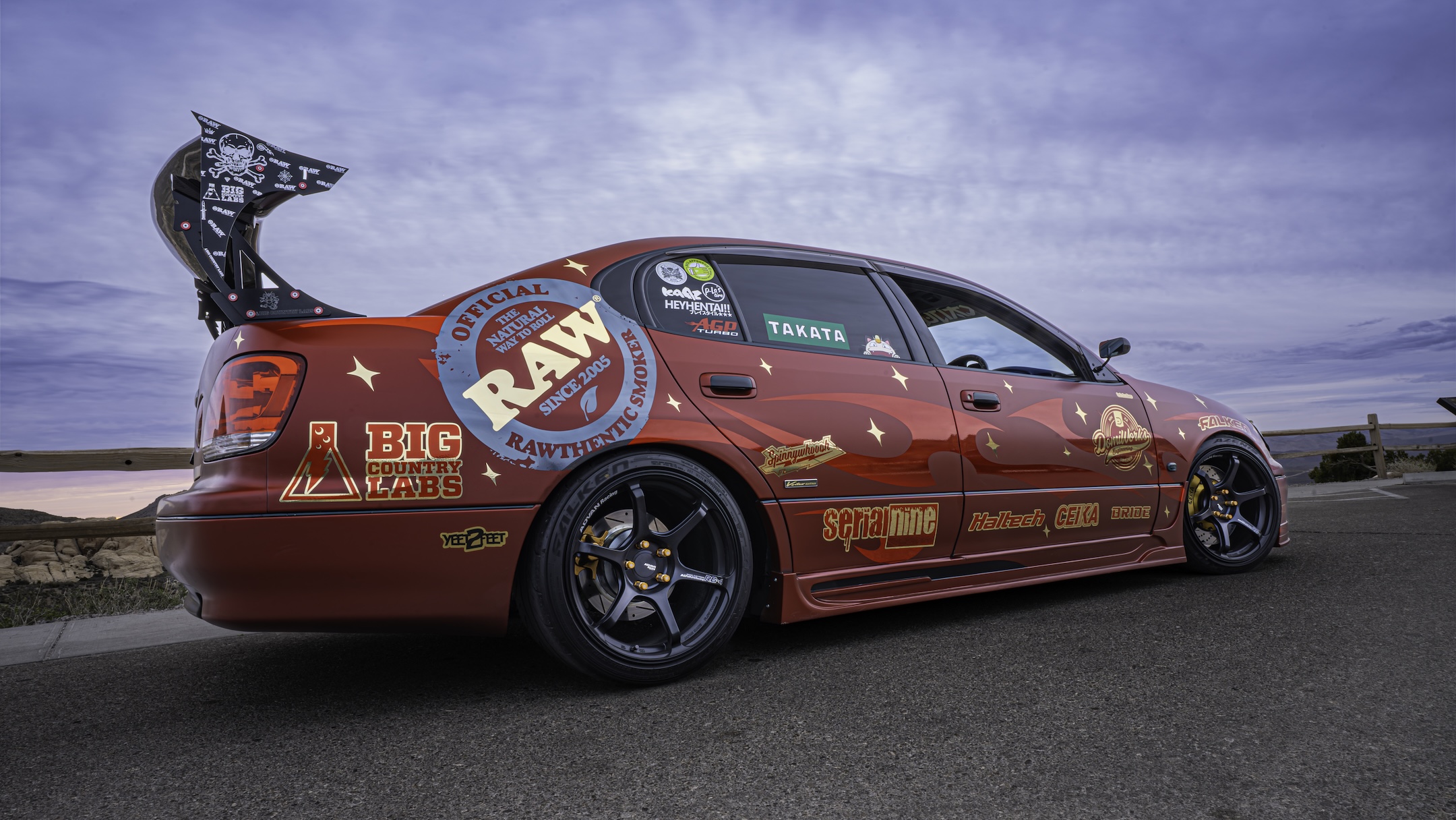 Rear three-quarter view of Chris Burch’s satin red 1997 Toyota Aristo showing the RAW livery, Big Country Labs wing, Advan RG4 wheels, Falken tires, and mountain backdrop.