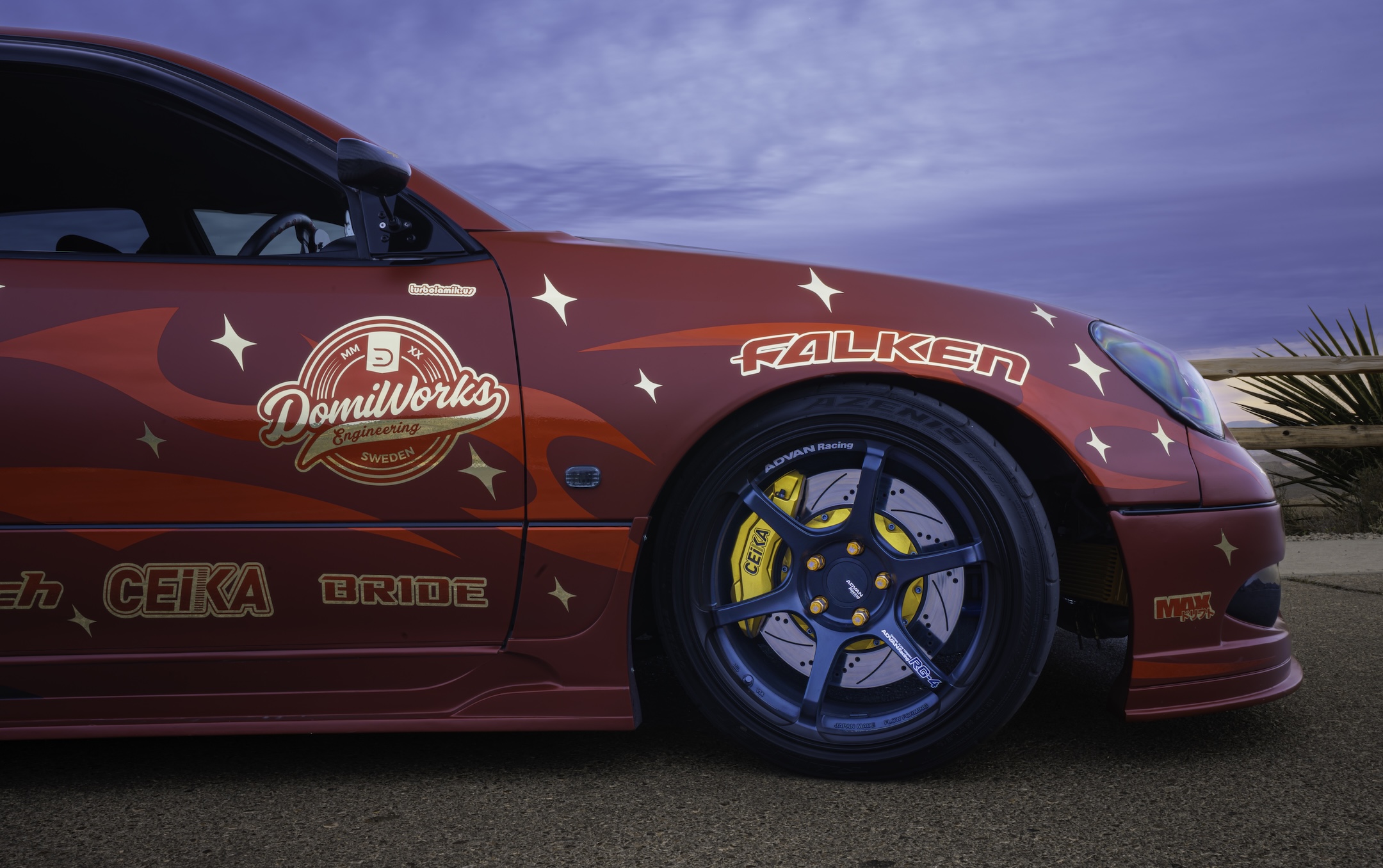 Close-up side detail of Chris Burch’s satin red 1997 Toyota Aristo showing the Advan RG4 wheel, Falken tire, CEIKA brake kit, Domiworks branding, and RAW-inspired side livery.