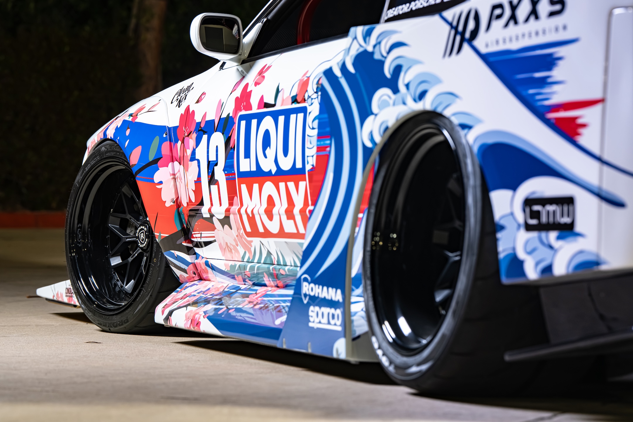 Close-up side detail of Osmond Seangsuwan’s 1990 Nissan Skyline GT-R showing the Liberty Walk LB-Kaido widebody, LIQUI MOLY livery, Rohana wheels, and Japanese-inspired wave and cherry blossom artwork.