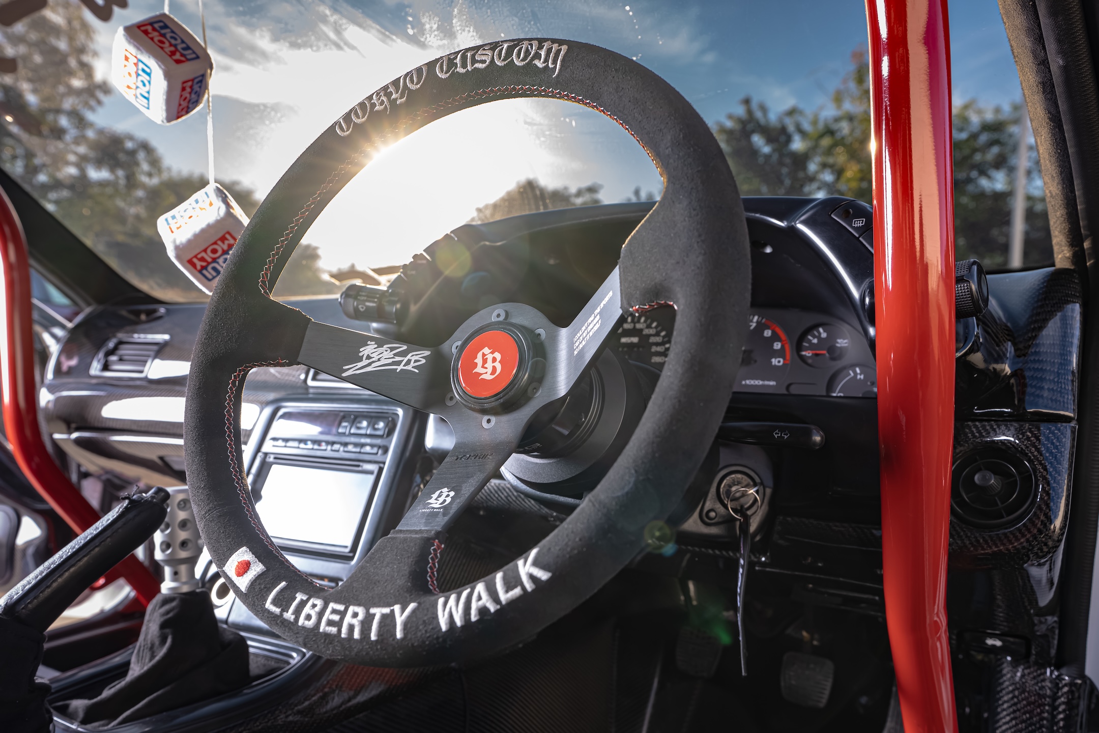 Close-up interior detail of Osmond Seangsuwan’s 1990 Nissan Skyline GT-R showing the Liberty Walk steering wheel, red Cusco roll cage, carbon interior trim, Alpine multimedia system, and right-hand-drive cockpit.