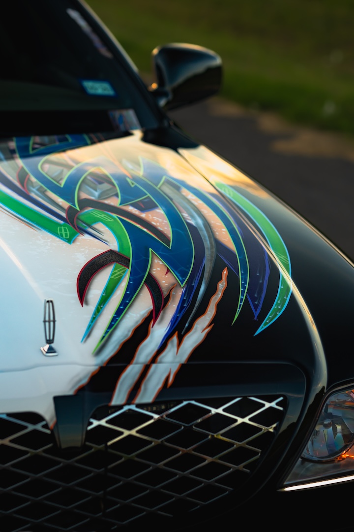 Close-up hood detail of Sam Benischek’s 2006 Lincoln Town Car showing the custom Two-Face graphic paint, Lincoln hood ornament, chrome grille, and split black-and-white front-end treatment.