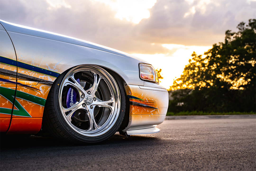 Close-up front wheel detail of Sam Benischek’s 2006 Lincoln Town Car showing the polished KRNC forged wheel, low air suspension stance, white Two-Face graphic paint, and front fender detail.