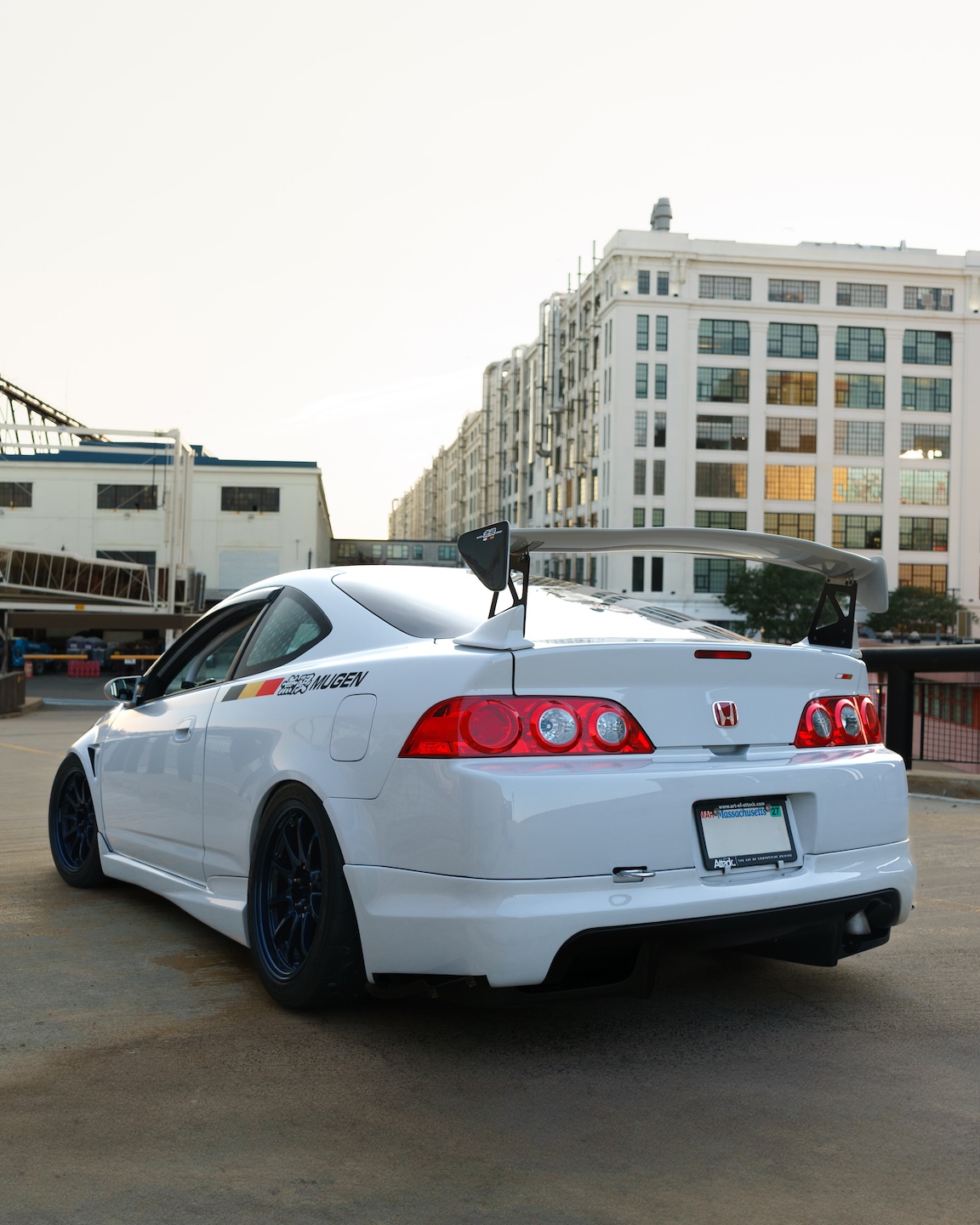 Rear three-quarter view of Jonathan Ortiz’s 2005 Acura RSX Type-S showing the Mugen wing, DC5R Kouki taillights, First Molding carbon diffuser, blue Volk Racing CE28N wheels, and Porsche PTS Fashion Grey paint.