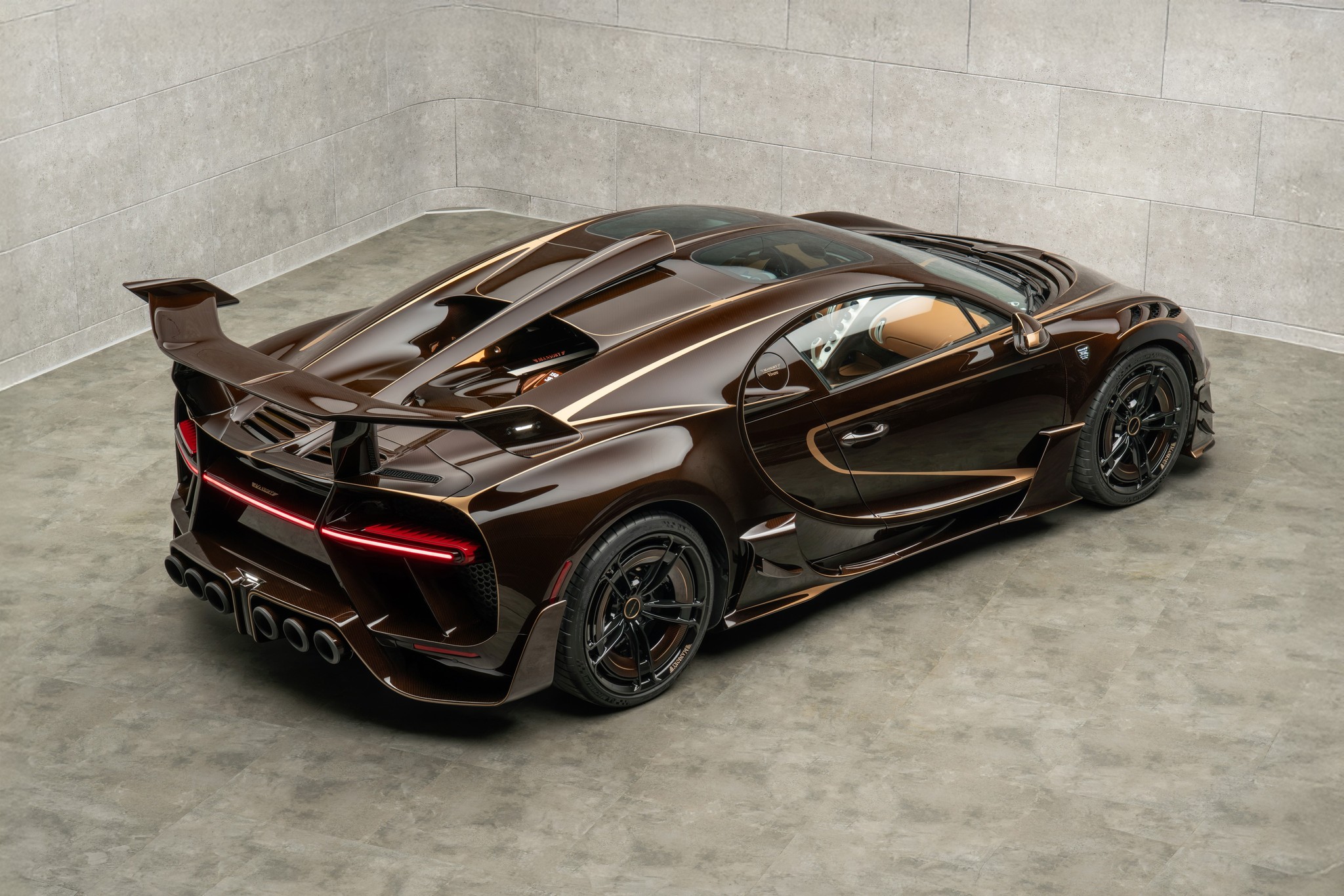 MANSORY Vivere Bugatti-based one-off shown from the rear three-quarter angle with brown exposed carbon bodywork, gold accents, black forged wheels, and large rear wing.