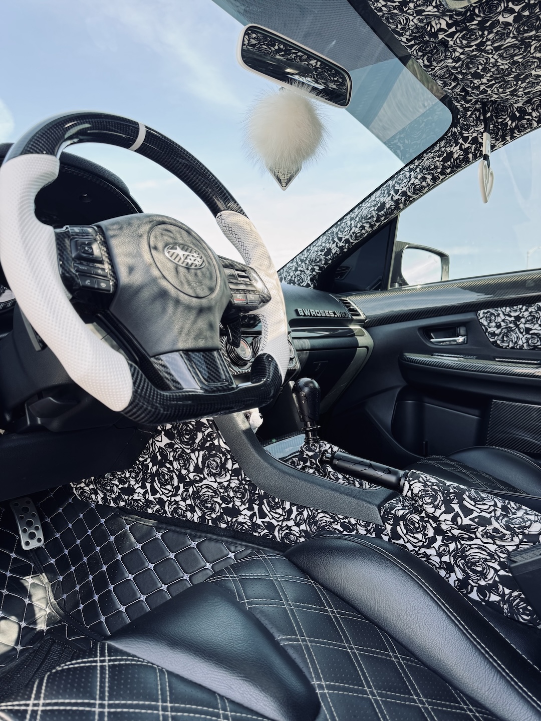 Cockpit detail of Melissa Pelletier’s 2017 Subaru WRX showing the carbon fiber steering wheel, rose-pattern interior trim, Billetworkz controls, custom door panels, and black-and-white cabin theme.