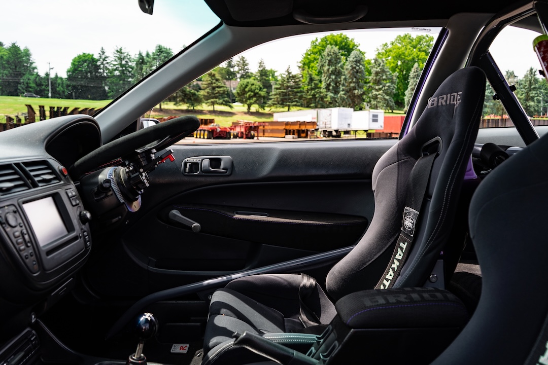 Right-hand-drive interior of Brandon Elste’s 1996 Honda Civic showing the Bride Stradia II seat, Takata harness, Mugen steering wheel, Works Bell flip hub, and EK4 OEM navigation.